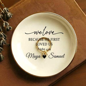 May include: A white ceramic ring dish with a gold rim, inscribed with the words "we love" and "BECAUSE HE FIRST LOVED US John 4:19". The names "Maya" and "Samuel" are also written on the dish. A gold ring rests in the center.
