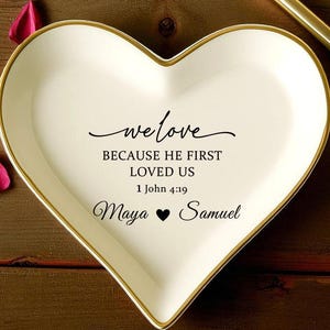 May include: A heart-shaped white ceramic dish with a gold rim, inscribed with the words "we love BECAUSE HE FIRST LOVED US 1 John 4:19 Maya ❤ Samuel." Two gold rings, a gold pen, and rose petals are also visible.