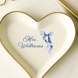 May include: Heart-shaped white ceramic trinket dish with gold trim. The dish features the name "Mrs. Williams" in blue script and a blue ribbon design. Two gold rings are visible near the dish.