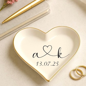 May include: A heart-shaped, off-white ceramic ring dish with gold trim. Black script reads "a & k" with a heart symbol and the date "13.07.25". Two gold rings sit beside the dish. A gold pen and a small pink flower are also present.