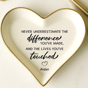 May include: A heart-shaped, white ceramic trinket dish with a gold rim. The dish features the text "Never underestimate the difference you've made, and the lives you've touched" with a heart and the name "Aiden". Two gold rings are visible.