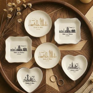 May include: A collection of personalized ring dishes in various shapes, including round, square, and heart-shaped. Each dish features a city skyline design with names and dates. The dishes are arranged on a wooden tray with a candle and dried flowers.