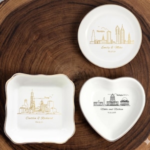 May include: Three white ceramic dishes with gold and black city skyline designs. One round dish features Philadelphia, another heart-shaped dish displays Washington D.C., and a square dish showcases Chicago. Each dish has names and dates.