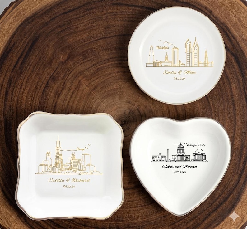 May include: Three white ceramic dishes with gold and black city skyline designs. One round dish features Philadelphia, another heart-shaped dish displays Washington D.C., and a square dish showcases Chicago. Each dish has names and dates.