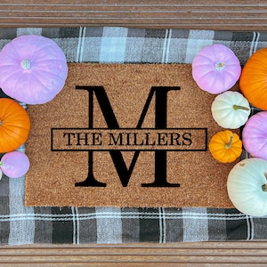 May include: A brown doormat with the text "THE MILLERS" and a large black "M" is surrounded by colorful pumpkins in shades of orange, white, and purple. The mat rests on a black and white checkered fabric.