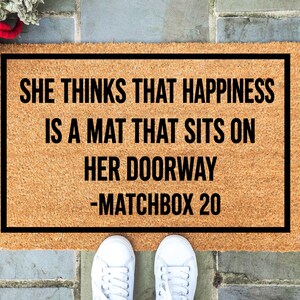 May include: A rectangular doormat with a black border and the text "SHE THINKS THAT HAPPINESS IS A MAT THAT SITS ON HER DOORWAY -MATCHBOX 20" in black. The mat is made of a light brown material. White sneakers are at the bottom.