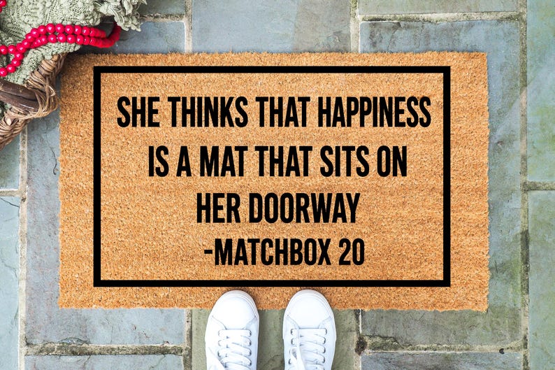 May include: A rectangular doormat with a black border and the text "SHE THINKS THAT HAPPINESS IS A MAT THAT SITS ON HER DOORWAY -MATCHBOX 20" in black. The mat is made of a light brown material. White sneakers are at the bottom.