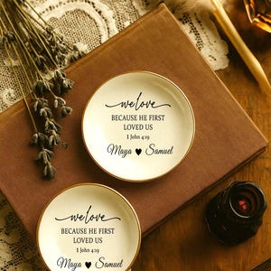 May include: Two small, round, white dishes with gold rims. Each dish has the words "we love" and "BECAUSE HE FIRST LOVED US 1 John 4:19 Maya &hearts; Samuel" in black script. The dishes rest on a brown surface.