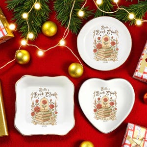 May include: White ceramic dishes in round, square, and heart shapes, each featuring a "Belle's Book Club" design with a rose, books, and the text "Tale as Old as Time." Gold trim edges the dishes, set against a red background with Christmas decorations.