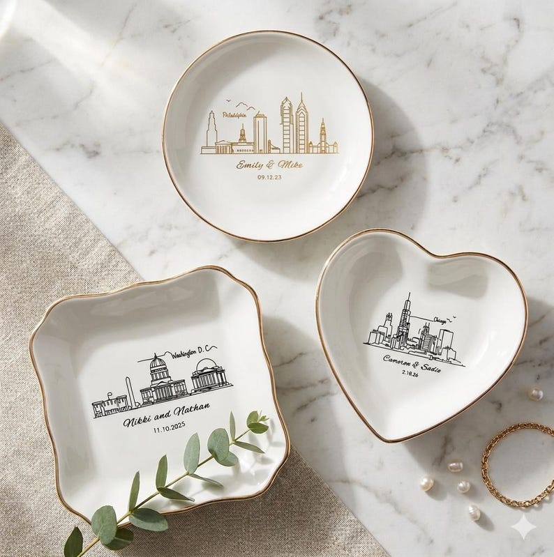 May include: Three white ceramic trinket dishes with gold rims. One round dish features a Philadelphia skyline, the second a Washington D.C. skyline, and the third a heart-shaped dish with a Chicago skyline. Each dish has personalized text.