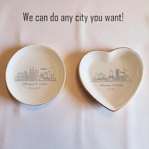 May include: Two white ceramic ring dishes with gold rims. One is round, the other heart-shaped. Each features a black skyline design of a city, with names and dates. Text above reads "We can do any city you want!"