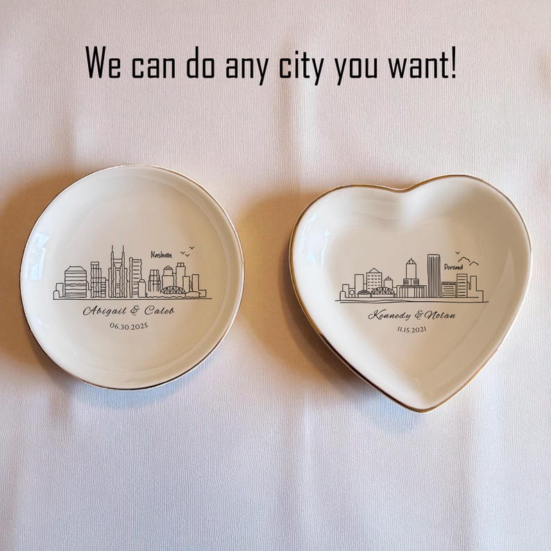 May include: Two white ceramic ring dishes with gold rims. One is round, the other heart-shaped. Each features a black skyline design of a city, with names and dates. Text above reads "We can do any city you want!"