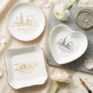 May include: Three white ceramic ring dishes with gold rims. One is round, one heart-shaped, and one square with scalloped edges. Each features a city skyline design and personalized text. A gold ring holder and flowers are also present.