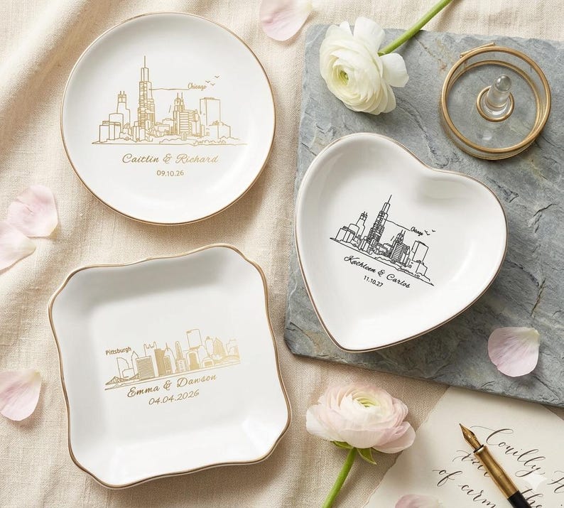 May include: Three white ceramic ring dishes with gold rims. One is round, one heart-shaped, and one square with scalloped edges. Each features a city skyline design and personalized text. A gold ring holder and flowers are also present.