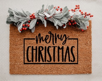 May include: A rectangular doormat with the words "merry CHRISTMAS" in black script. The mat is brown and has a decorative border of gray leaves and red berries across the top. The mat is designed for the holiday season.