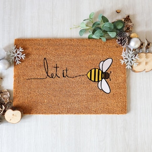 May include: A rectangular doormat made of brown coir fiber. The mat features the words "let it" in a handwritten font, with a cartoon bee graphic. The bee has yellow and black stripes and white wings. Decorative elements surround the mat.