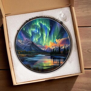 May include: Round wall art featuring a vibrant depiction of the Aurora Borealis over a mountain range and a reflective lake. The artwork is framed in black and presented in a cardboard box with a suction cup attached.