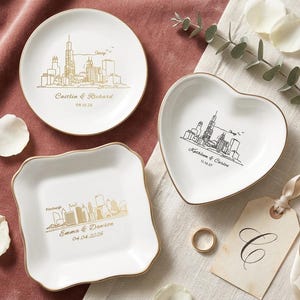 May include: Three white ceramic dishes with gold rims. One round, one heart-shaped, and one square with a wavy edge. Each features a city skyline design and personalized text. A gold ring and a tag with a letter are also present.