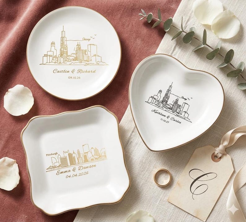 May include: Three white ceramic dishes with gold rims. One round, one heart-shaped, and one square with a wavy edge. Each features a city skyline design and personalized text. A gold ring and a tag with a letter are also present.