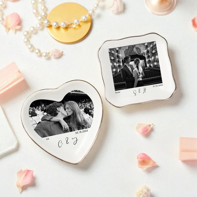 May include: Two white ceramic ring dishes with gold trim. One is heart-shaped, the other is square with scalloped edges. Both feature black and white photos of couples, with dates and initials. Decorative items.