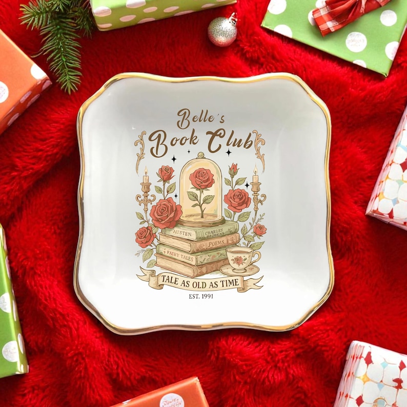 May include: A white, square trinket dish with a gold rim, featuring the text "Belle's Book Club" and a design of a rose in a glass dome, books, and roses. The phrase "Tale as Old as Time" is also present.