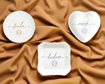 May include: Three white ceramic trinket dishes with gold or silver trim. One round dish has "linda" in gold script, a heart-shaped dish has "mark" in blue script, and a square dish has "barbara" in black script. Each dish features a volleyball graphic.