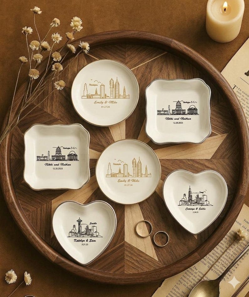 May include: A collection of personalized ring dishes in various shapes, including round, square, and heart-shaped. Each dish features a city skyline design with names and dates. The dishes are arranged on a wooden tray with a candle and dried flowers.