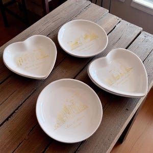 May include: Four white ceramic dishes with gold skyline designs and text. Two are heart-shaped, and two are round. The dishes have a gold rim and are arranged on a rustic wooden surface. Each dish has a different city skyline.