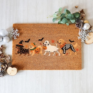 May include: A rectangular tan coir doormat featuring a printed graphic of four dogs in Halloween costumes. The dogs include a dachshund in a witch hat, a fluffy dog in a pumpkin suit, a dalmatian as a ghost, and a golden retriever in a skeleton suit.