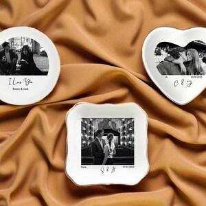 May include: Three white ceramic dishes with gold trim, each featuring a black and white photograph. One is round with "I Love You" text, another heart-shaped, and the third square. The dishes are on a gold fabric.