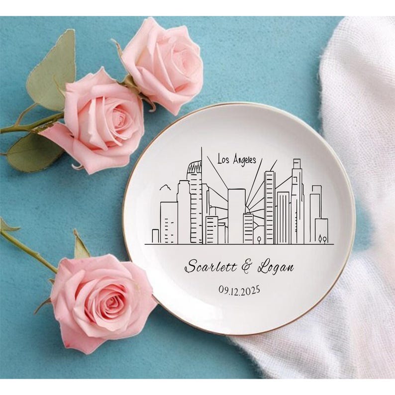 May include: A white ceramic plate with a gold rim features a black line drawing of the Los Angeles skyline. The plate is personalized with the names "Scarlett & Logan" and the date "09.12.2025". Pink roses are arranged around the plate.