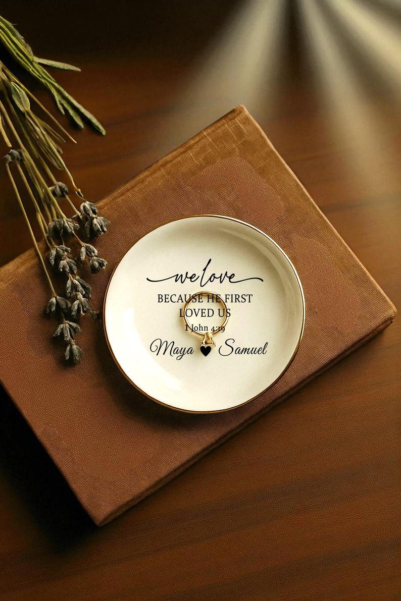 May include: A white ceramic ring dish with a gold rim, inscribed with the words "we love" and "BECAUSE HE FIRST LOVED US John 4:19". The names "Maya" and "Samuel" are also written on the dish. A gold ring rests in the center.
