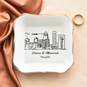 May include: A white ceramic trinket dish with a gold rim, featuring a black line drawing of the Louisville skyline. The dish is personalized with the names "Claire & Maverick" and the date "09.16.2025". A gold ring rests on the side.