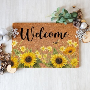 May include: A rectangular doormat with a brown coir surface, featuring the word "Welcome" in black script. The mat is decorated with yellow sunflowers, daffodils, and a bee. Decorative elements like snowflakes and pinecones surround the mat.