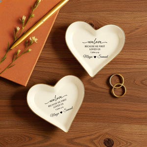 May include: Two heart-shaped, white ceramic ring dishes with gold trim. Each dish has the text "we love BECAUSE HE FIRST LOVED US 1 John 4:39 Maya ❤ Samuel". Gold rings and a pen are also visible.
