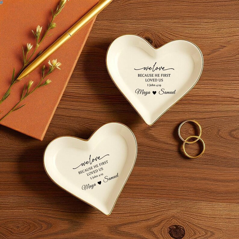 May include: Two heart-shaped, white ceramic ring dishes with gold trim. Each dish has the text "we love BECAUSE HE FIRST LOVED US 1 John 4:39 Maya ❤ Samuel". Gold rings and a pen are also visible.