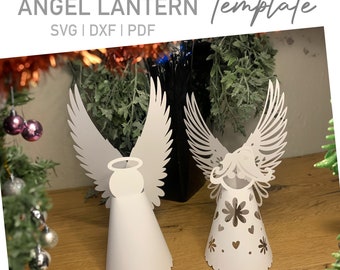Può includere: Due modelli di lanterne di angelo di carta bianca. L'angelo a sinistra ha un'aureola e ali semplici. L'angelo a destra ha un'aureola, ali e un viso più dettagliati con fiori e cuori ritagliati.