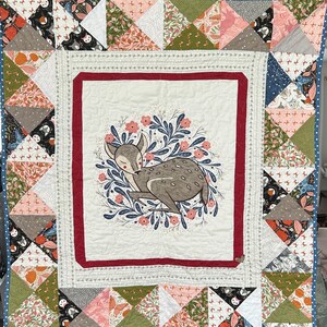 May include: A handmade quilt with a central image of a sleeping deer surrounded by flowers. The quilt has a white background with a red border, and is framed by colourful patchwork triangles in various patterns and colours.