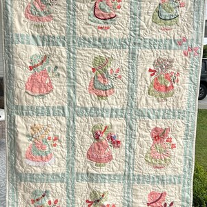 May include: Handmade quilt with appliqued figures of girls in bonnets and aprons, holding flowers. The quilt has a cream background with light blue and white striped borders. The figures are in pink, green, and red fabrics.