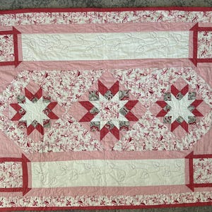 May include: A pink and red quilt with a floral pattern and a star design in the centre. The quilt is quilted with white thread and has a white border.