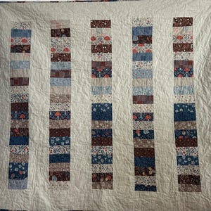 May include: A quilt featuring five vertical columns of patchwork fabric strips separated by solid cream-coloured fabric. The strips display various patterns including mushrooms, floral motifs, and geometric designs in shades of blue, brown, and red.