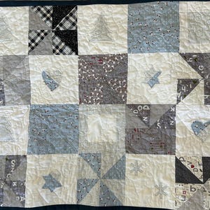 May include: A white quilt with a blue and grey patchwork design. The quilt is made up of squares and triangles, and features various patterns, including snowflakes, stars, and hearts. The quilt is edged with a dark blue border.