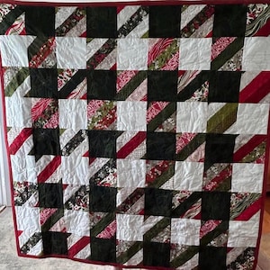 May include: A handmade quilt with a patchwork design. The quilt features a repeating pattern of squares in white, black, and various patterned fabrics in red, green, and floral prints. The quilt is bordered with a solid red fabric.