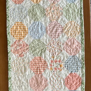 May include: A rectangular quilted table runner with a patchwork design. The runner features various pastel-coloured octagons with floral and gingham patterns. A green floral border frames the runner, which is placed on a wooden surface.