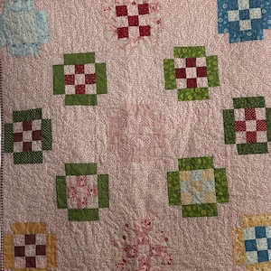 May include: A patchwork quilt featuring a light pink background with a delicate white floral pattern. The quilt is decorated with various square blocks in shades of blue, green, red, and yellow, each containing a smaller nine-patch grid design.