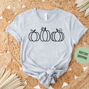 以下が含まれることがあります： グレーのTシャツに、黒い輪郭で描かれた3つのカボチャがプリントされています。カボチャは一列に並んでおり、茎と葉が付いています。