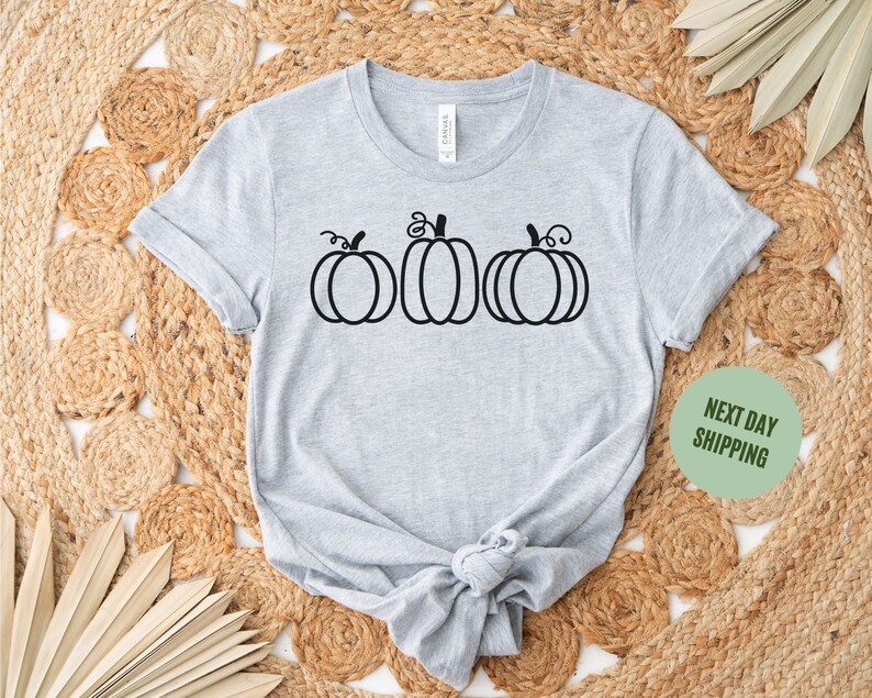 以下が含まれることがあります： グレーのTシャツに、黒い輪郭で描かれた3つのカボチャがプリントされています。カボチャは一列に並んでおり、茎と葉が付いています。