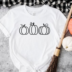 以下が含まれることがあります： 前面に黒い輪郭で3つのカボチャが描かれた白いTシャツ。カボチャは茎と葉でスタイリッシュに描かれています