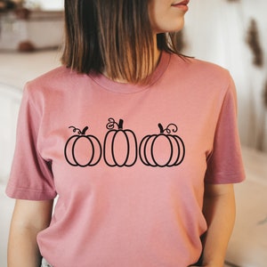 以下が含まれることがあります： 前面に黒い輪郭の3つのカボチャが描かれたピンクのTシャツです。