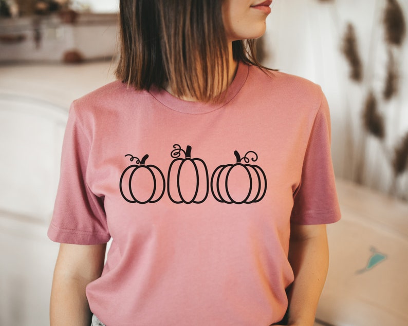 以下が含まれることがあります： 前面に黒い輪郭の3つのカボチャが描かれたピンクのTシャツです。